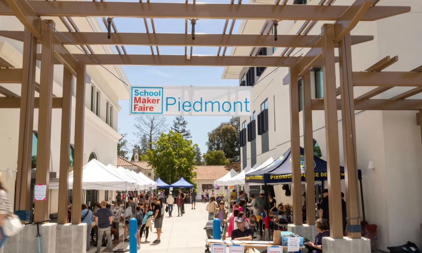 Visitors walk under a wooden pergola beneath a School Maker Faire Piedmont banner, with white exhibitor tents lining the sunny plaza on both sides.