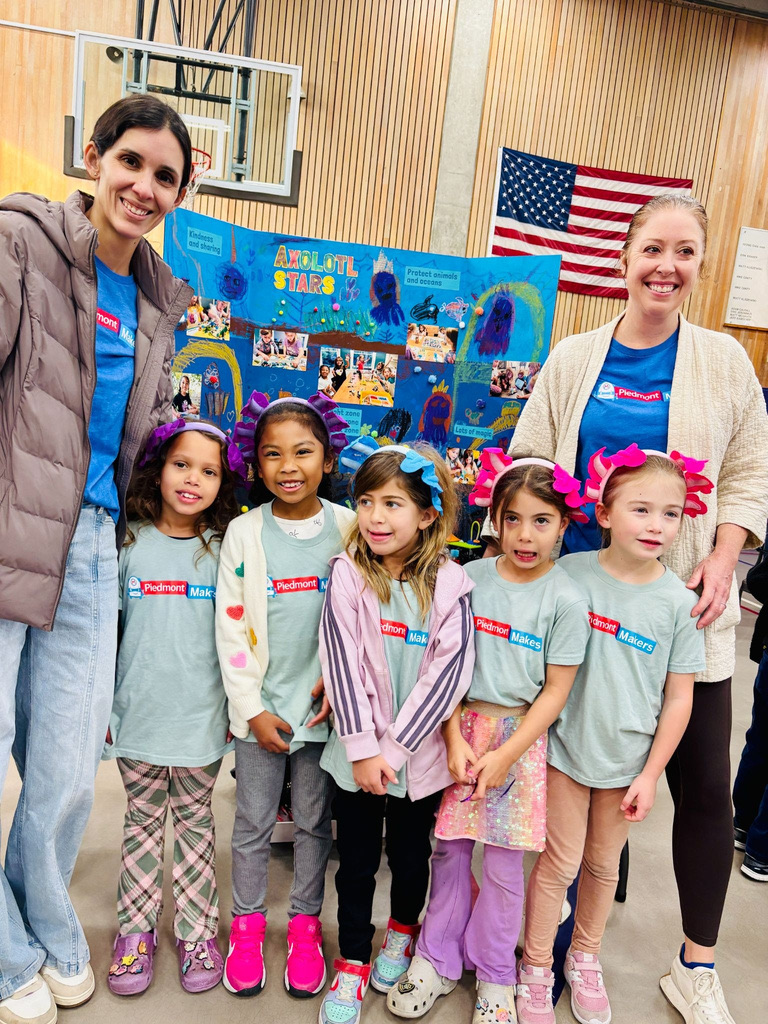 Five young LEGO League Explore teammates in Piedmont Makers shirts and colorful headbands stand with their two adult coaches in front of a hand-decorated project board reading Axolotl Stars.