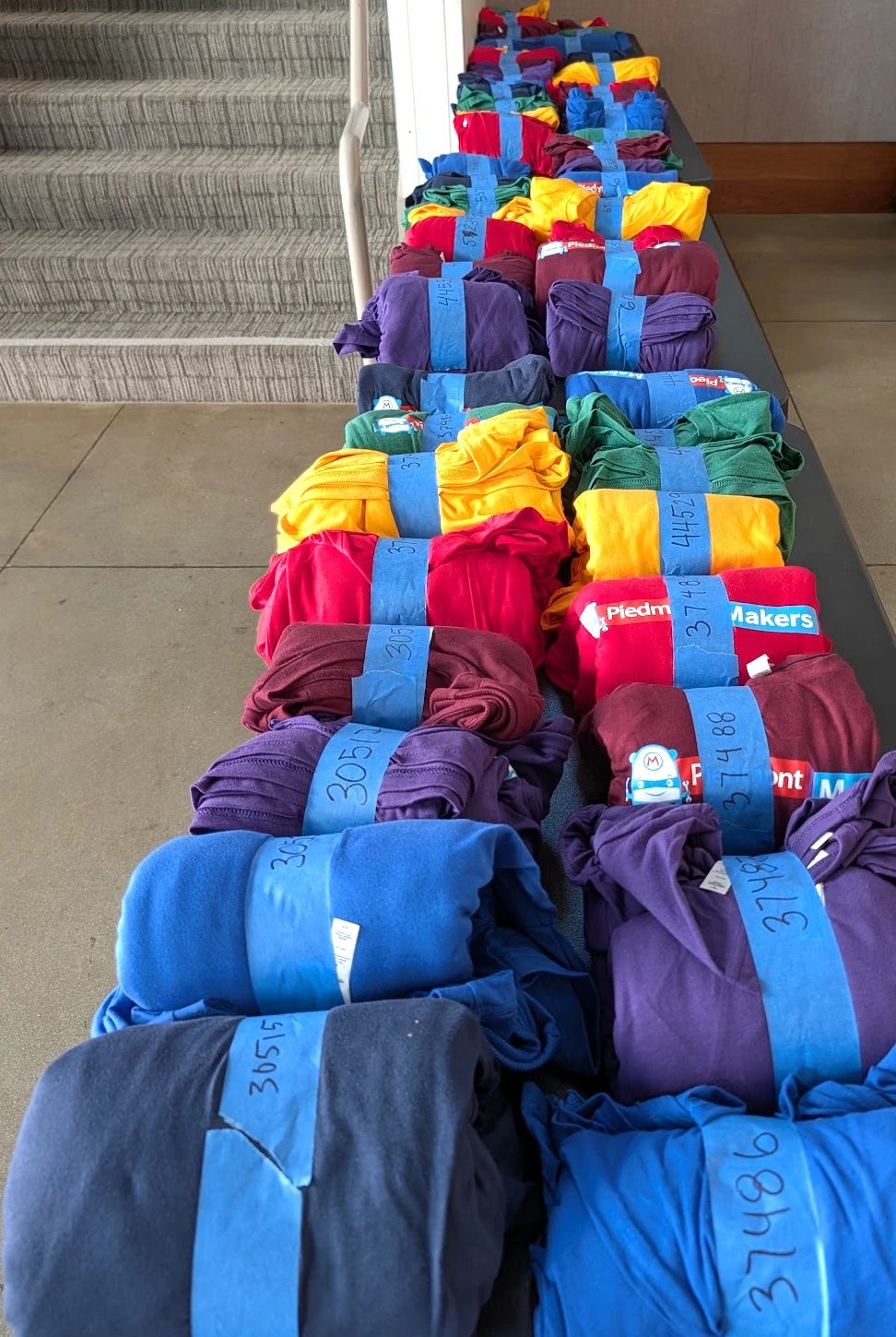 Dozens of folded team shirts in red, purple, gold, green, and blue, each bundle wrapped in blue tape and marked with a team number, lined up along a bench for pickup.