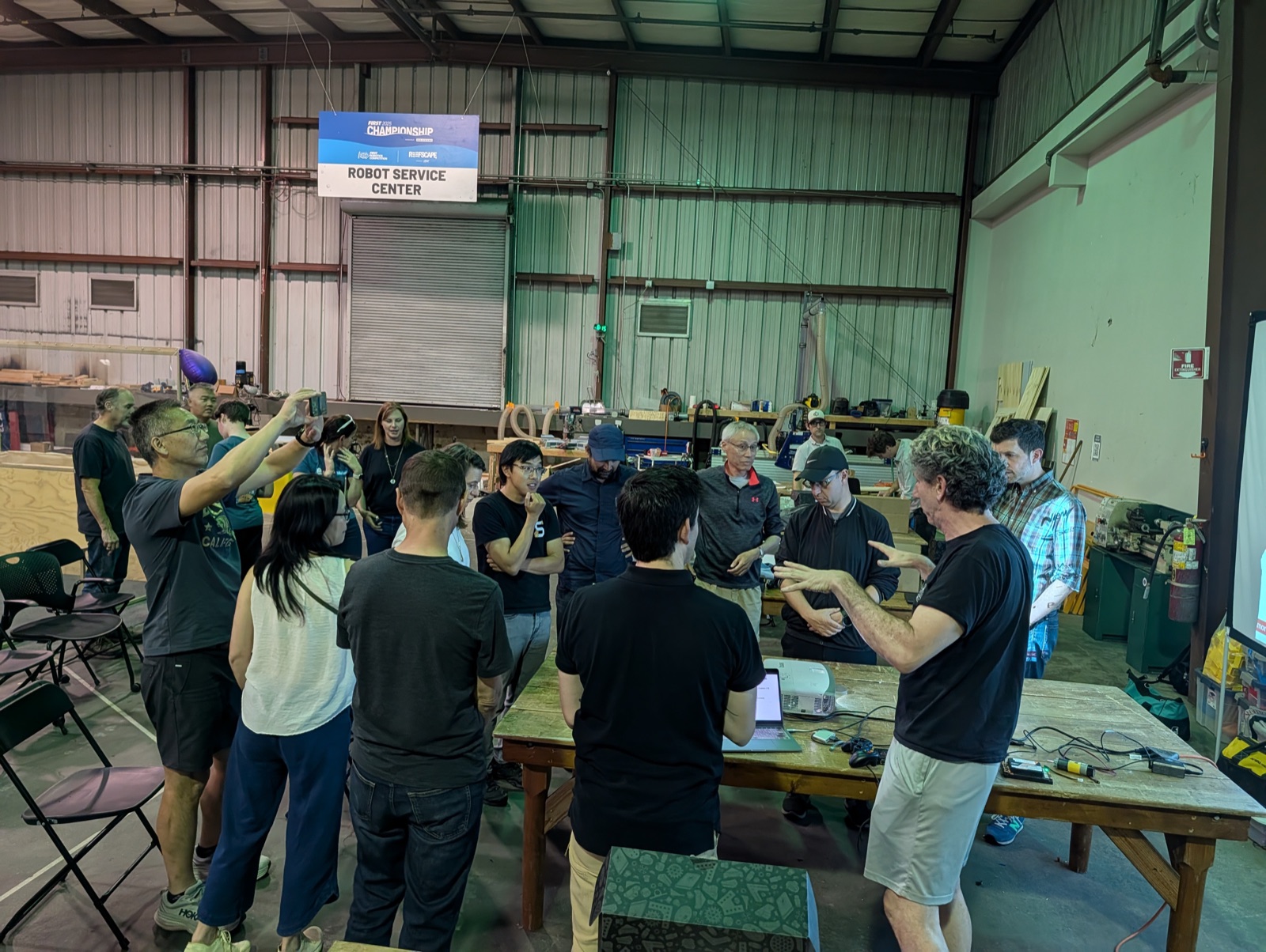 About fifteen adults standing in a circle around a wooden workbench in a warehouse, watching an instructor gesture over an open laptop, under a hanging FIRST Championship Robot Service Center sign.