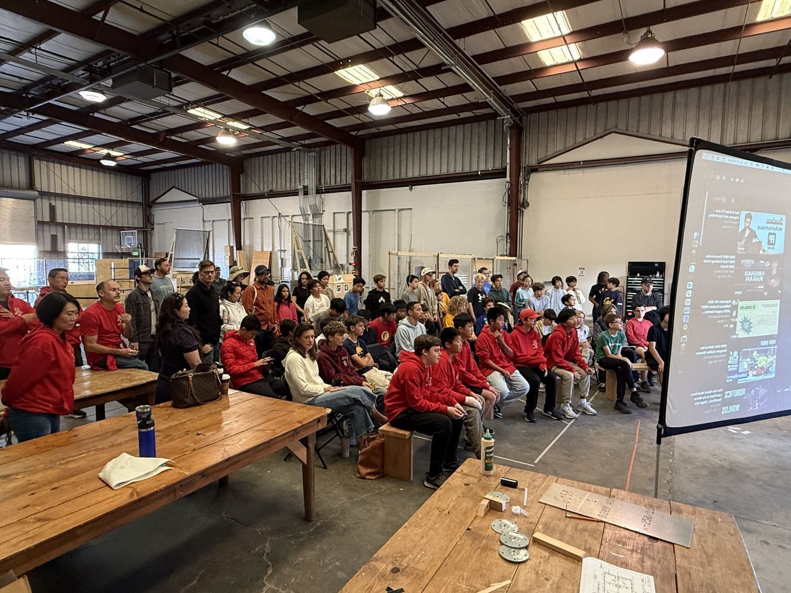 Dozens of students, many in red team hoodies, seated on benches in a warehouse maker space with parents and coaches standing behind them, all watching the FIRST Tech Challenge kickoff broadcast on a large projection screen.