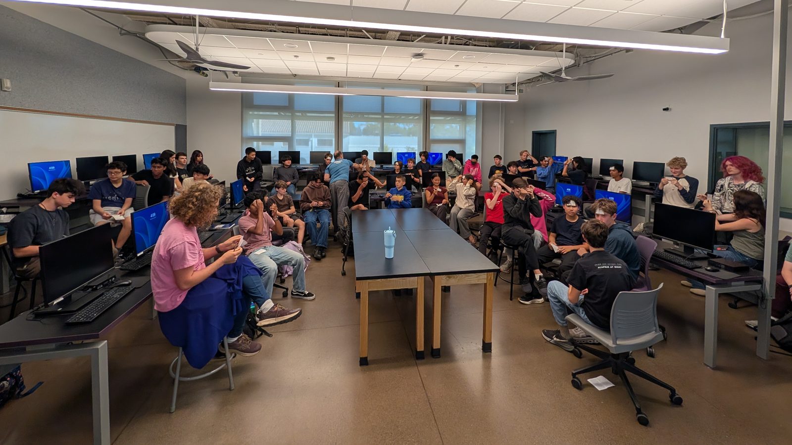 Dozens of high school students seated around the edges of a computer lab, several holding paper cards up to their foreheads during an icebreaker game.