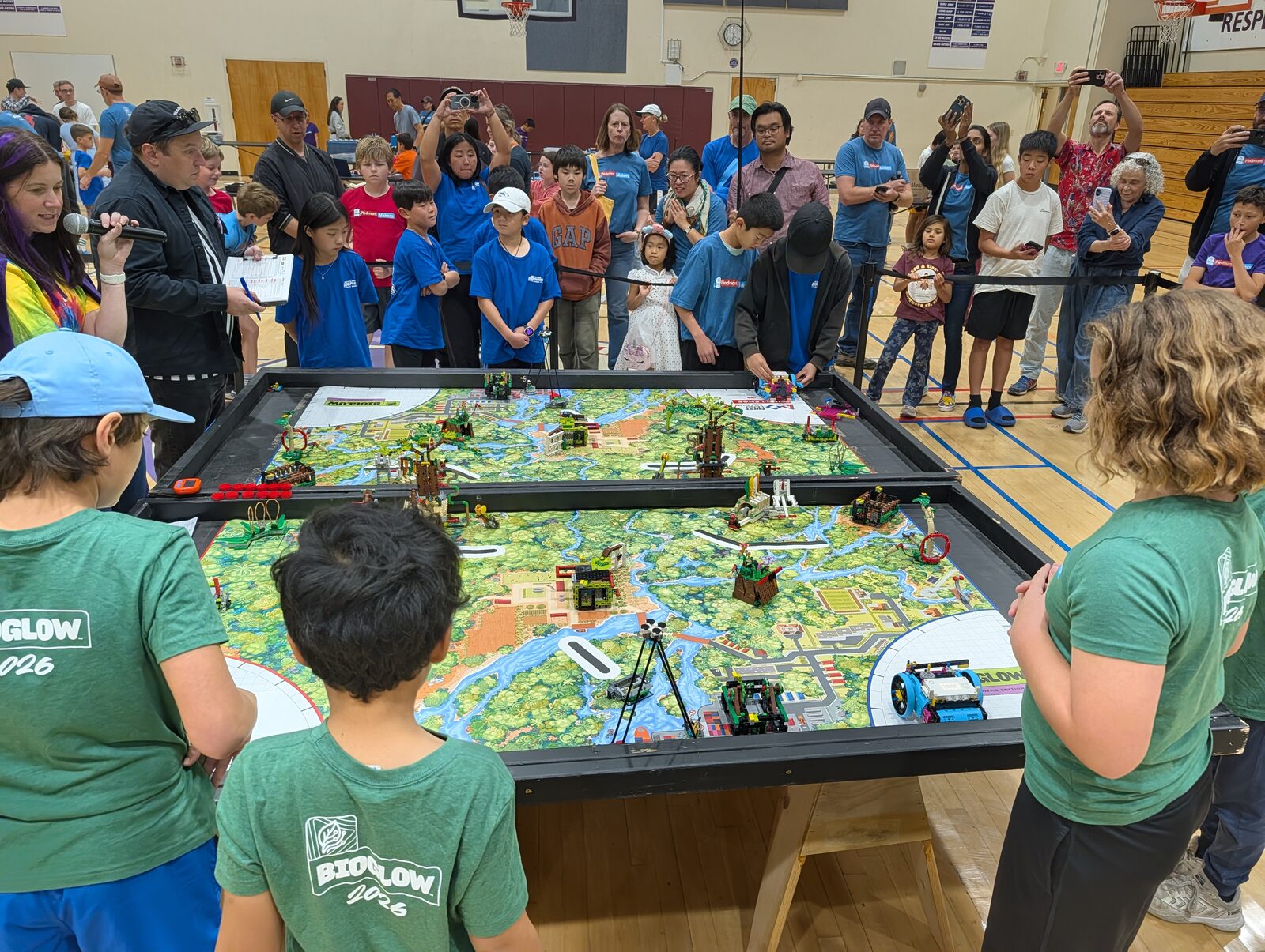 Two teams in green and blue shirts on opposite sides of a pair of game tables covered with LEGO mission models, while a large crowd of kids and parents watches and an announcer speaks into a microphone at the left.