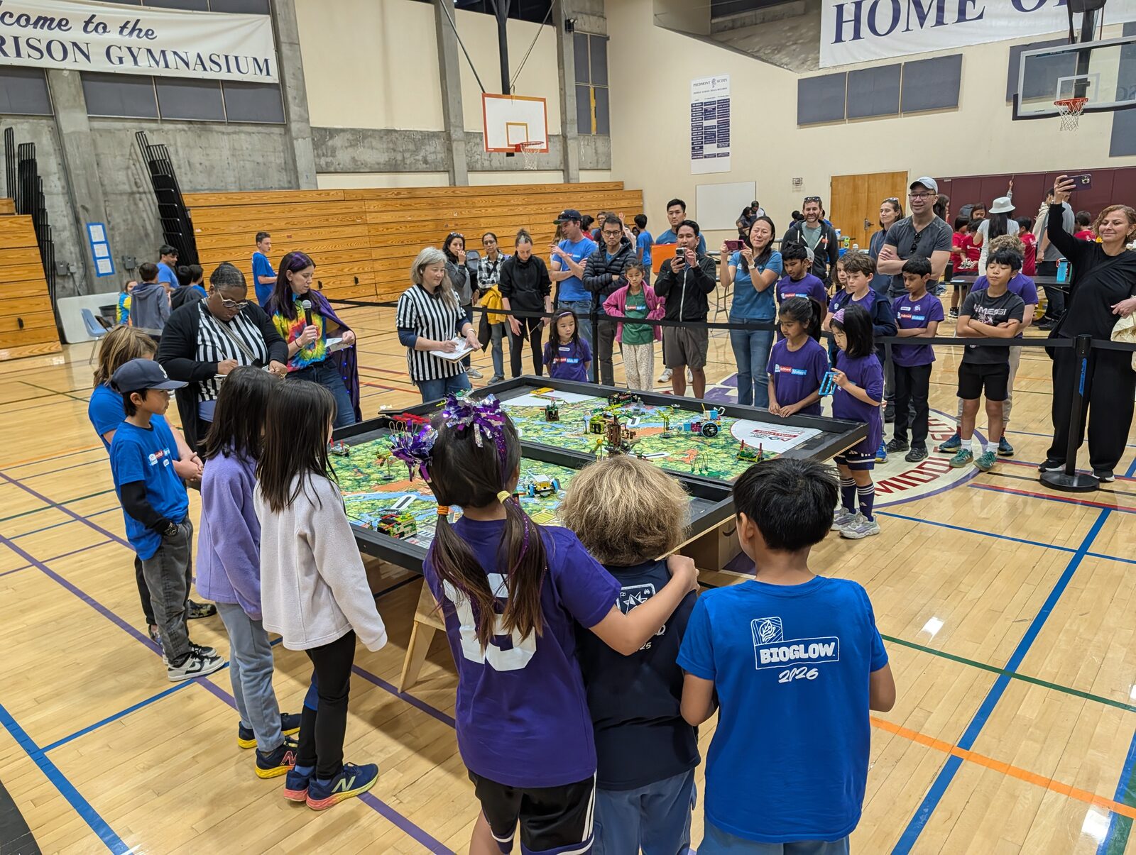 LEGO League Challenge teams in matching shirts stand around a robot game table in the Morrison Gym while two referees in striped shirts score, an announcer with a microphone calls the round, and parents behind a rope line film on their phones.