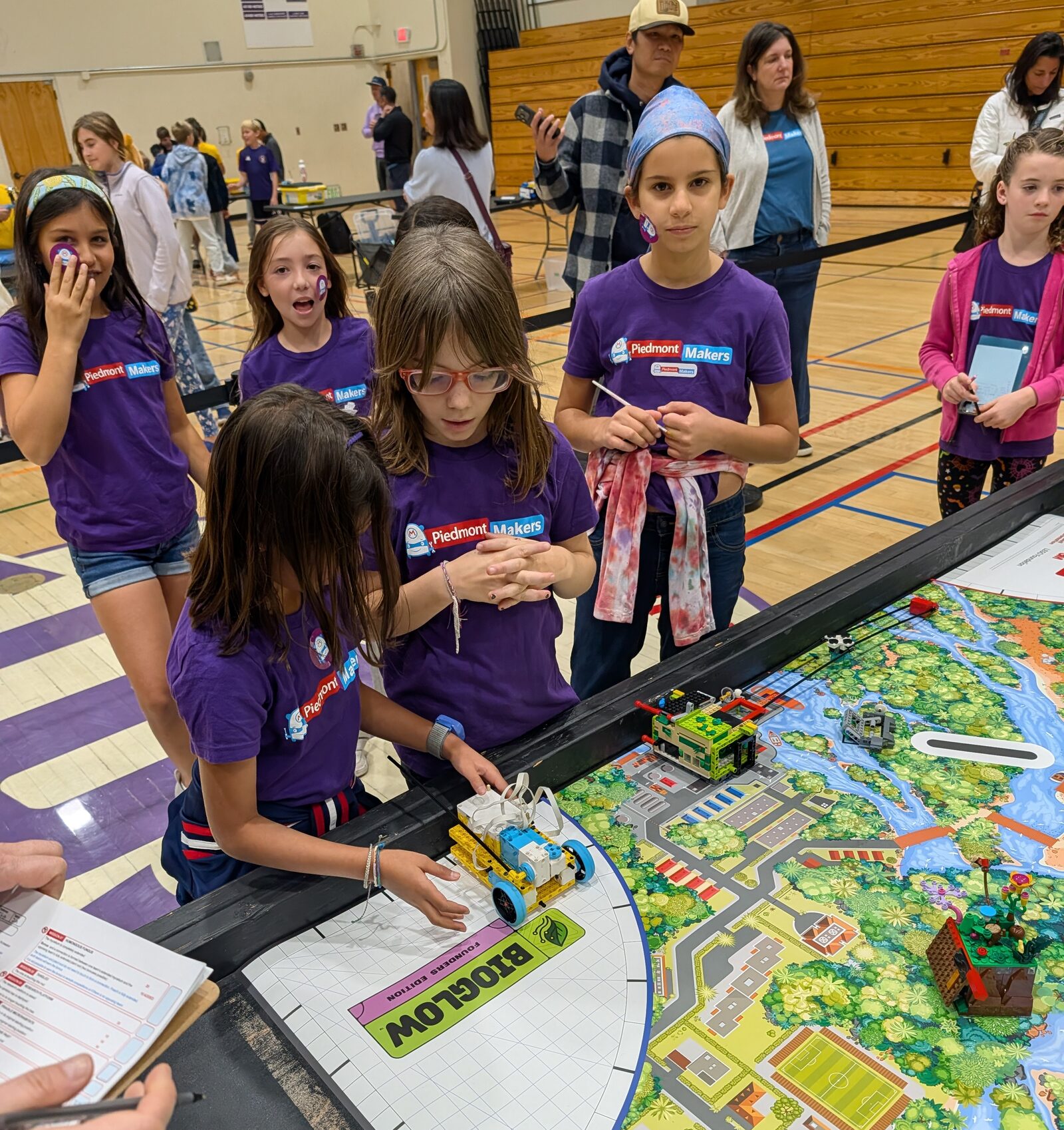 A team of girls in purple Piedmont Makers shirts at the edge of a game table. One lines up a small LEGO robot in the launch area beside the BIOGLOW logo while teammates watch and a referee holds a scoresheet.
