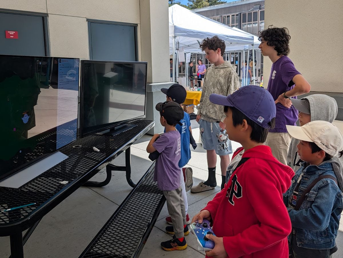 A cluster of kids gathered around two monitors on an outdoor lunch table, one holding a game controller, while older students look on.