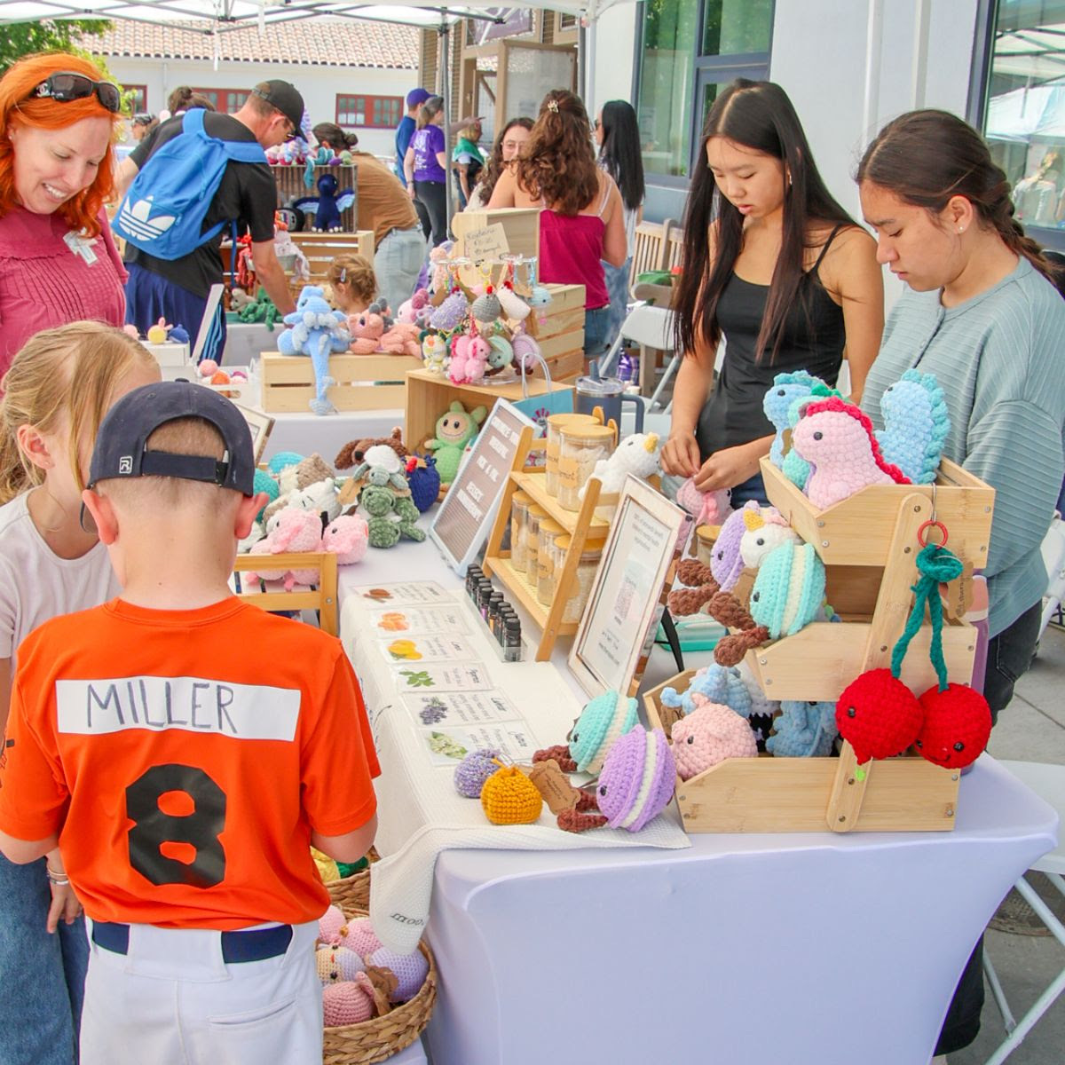 Two teenage makers tend a booth full of pastel crocheted animals while younger kids and parents browse the table.