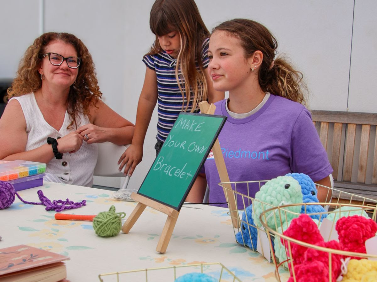 A young volunteer in a purple Piedmont Makers shirt sits with an adult and a younger child at a make-your-own-bracelet table topped with colorful yarn and crocheted creatures.