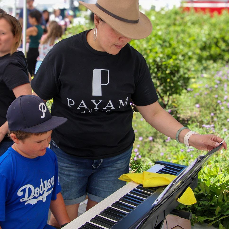 A Payam Music instructor in a wide-brim hat guides a young boy at an electronic keyboard set up among garden greenery.