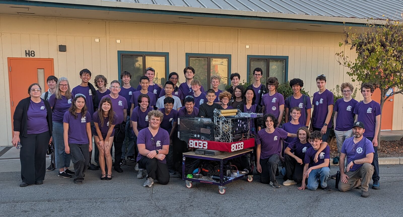 About forty Highlander Robotics students and mentors in purple Team 8033 shirts posed outside a portable classroom around their robot, which sits on a wheeled cart with red 8033 bumpers.