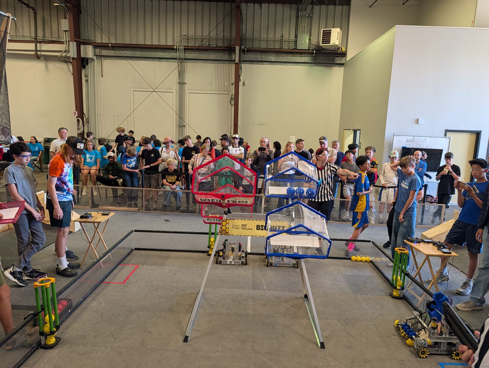 An FTC match in progress in a warehouse: small metal robots on a gray tile field with red and blue hive-shaped goals, students at driver stations on both sides, a referee pointing, and a crowd of families watching from behind the field wall.