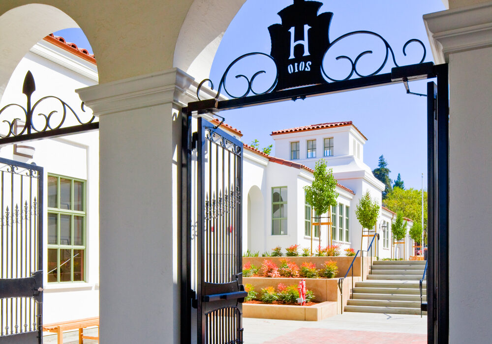 The entrance gate at Havens Elementary, a black iron archway topped with a crest bearing the letter H, opening onto a white stucco school building with red tile roofs, planters, and wide outdoor steps.