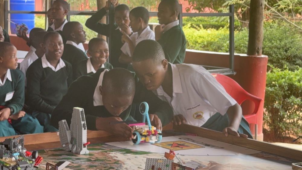 Two SEGA students lean over a FIRST LEGO League Unearthed mission mat, lining up their LEGO SPIKE robot while classmates in school uniforms watch from behind.