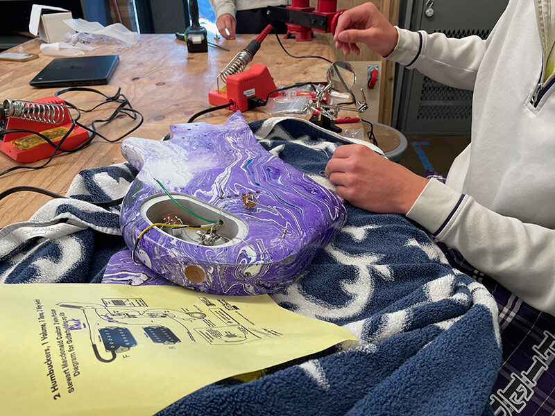 A Piedmont High student solders the wiring inside a marbled purple electric guitar body, with soldering irons and a printed wiring diagram on the workbench.