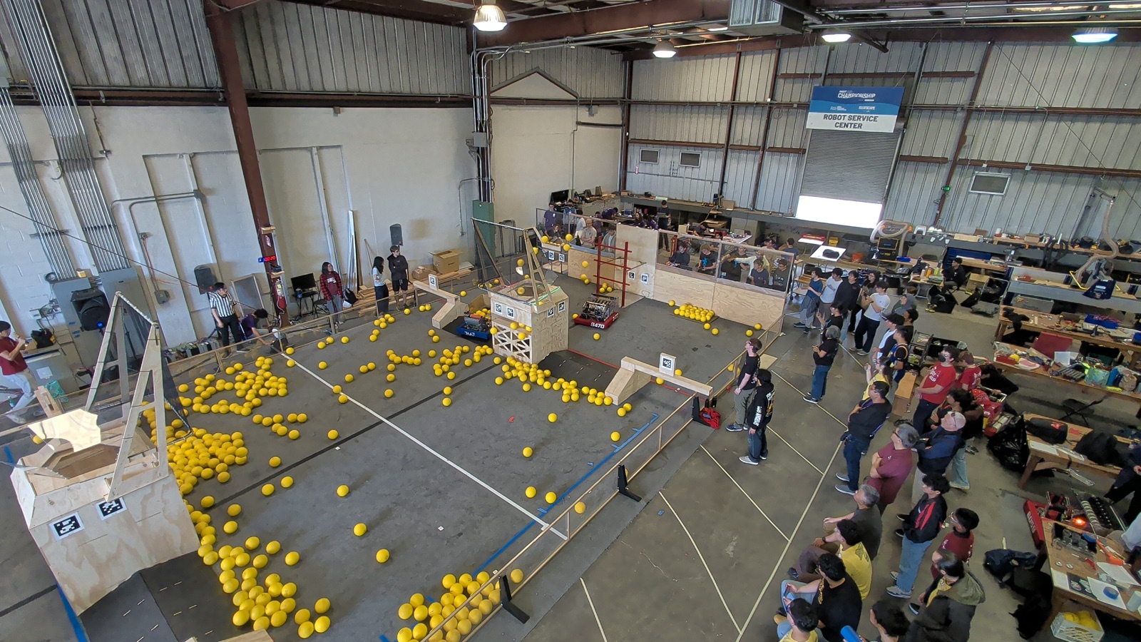 Overhead view of the 10th Street practice field — a regulation-size FRC competition area with hundreds of yellow game pieces scattered across the floor, robots on the field, and the team and spectators around the edges.