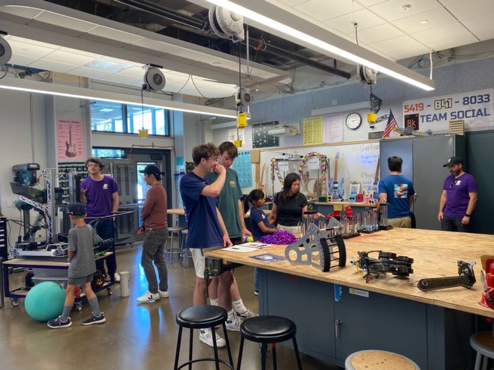 Interior of the Mary G. Ross Engineering Lab at Piedmont High School — high schoolers in purple shirts working at tall plywood benches around an FRC robot frame, with team banners, the Team Social board, and FRC parts visible in the background.