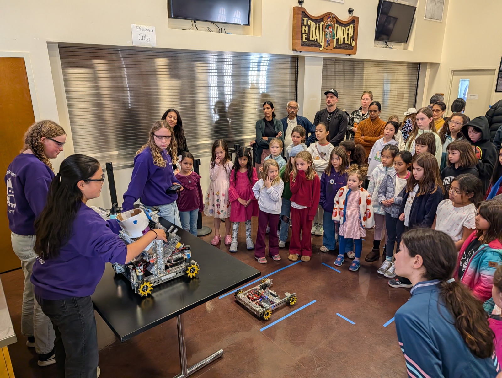 High school students from FRC Team 8033 in purple Piedmont Makers sweatshirts demonstrating a robot to a crowd of younger girls.