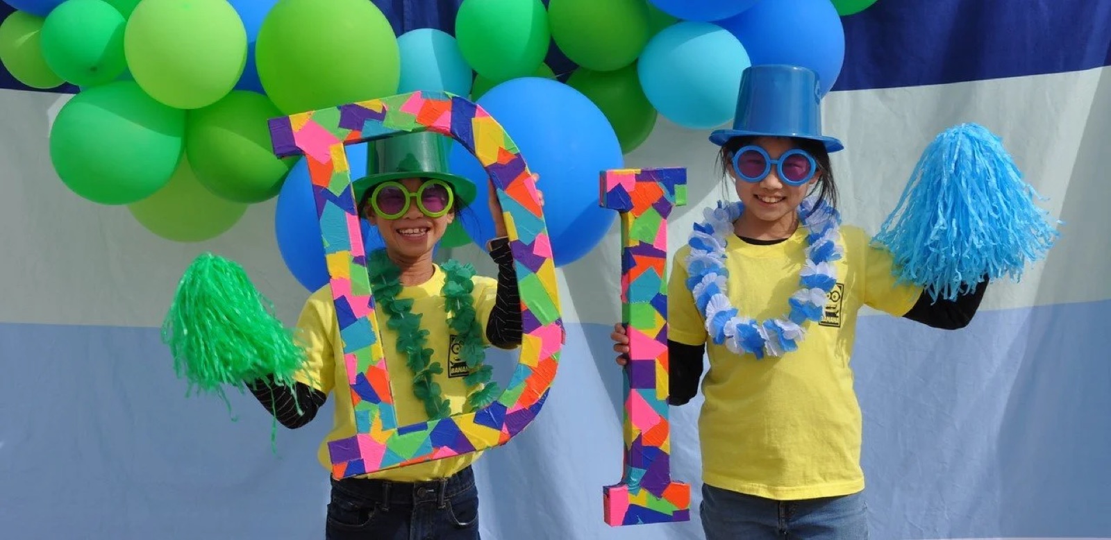 Two kids in bright costume hats and pom-poms holding handmade letters spelling 'DI' in front of a green and blue balloon arch.