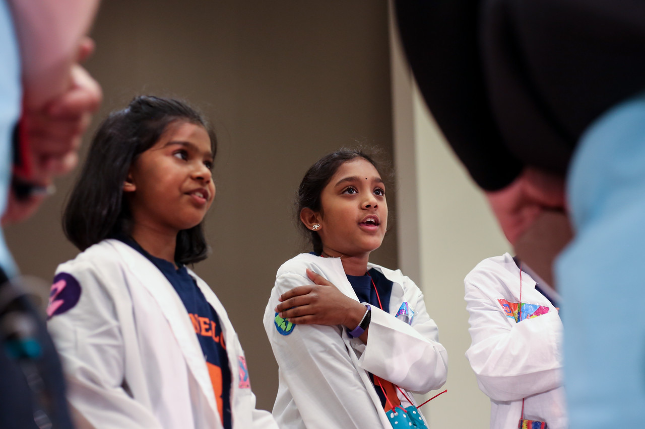 Three young girls in white lab coats decorated with handmade patches, watching attentively at a Destination Imagination tournament.