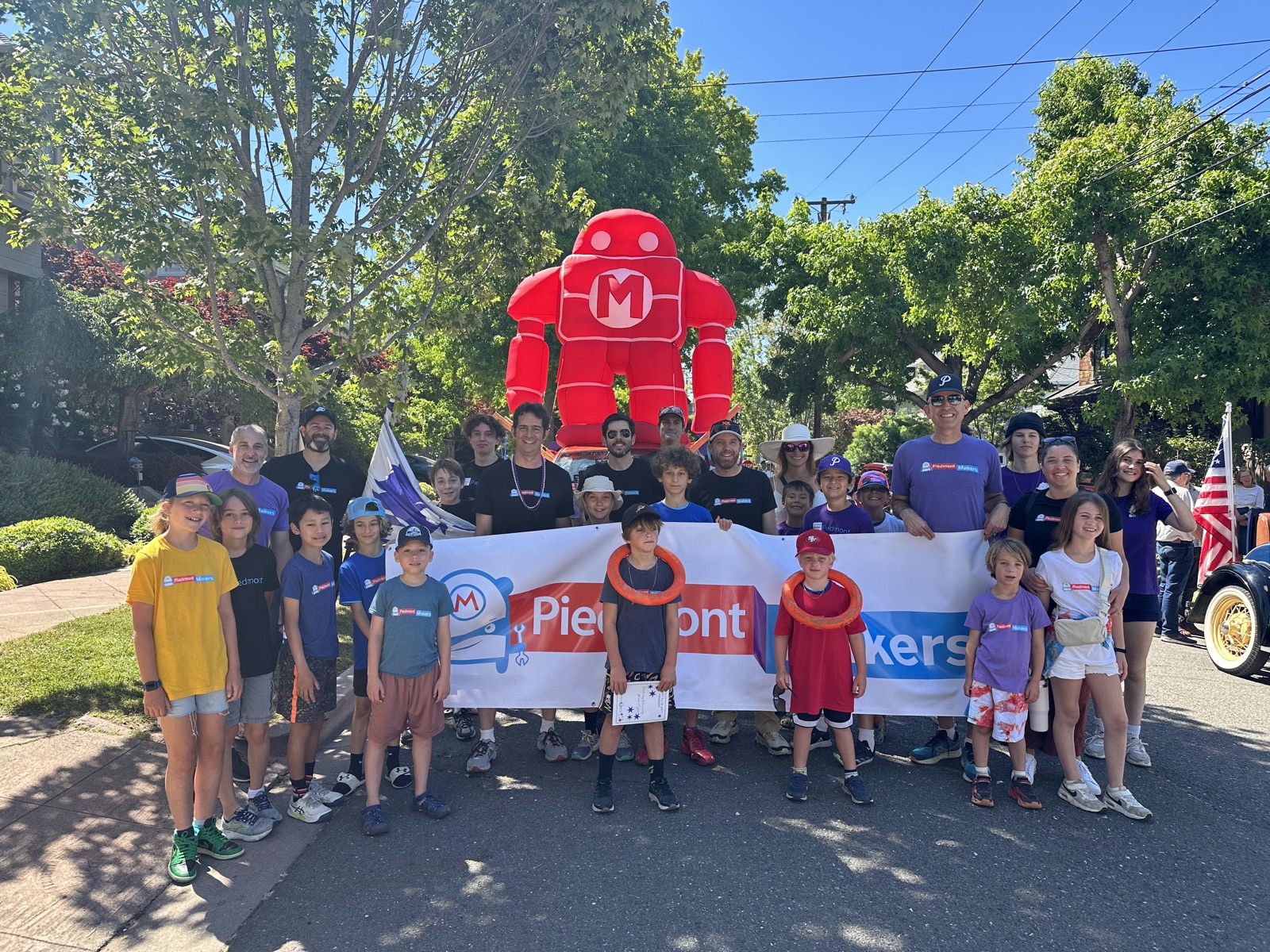 A large group of Piedmont Makers kids and parents in matching shirts posing with the Piedmont Makers banner in front of a 15-foot inflatable red Makey robot on Highland Avenue.