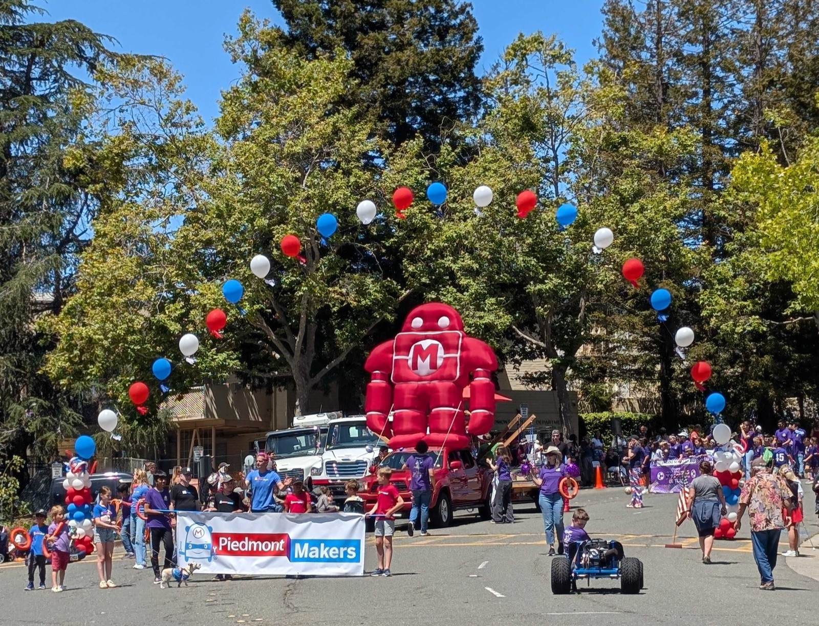 The giant red inflatable Makey rising above the 4th of July Parade — kids in purple Piedmont Makers shirts hold the banner below, red-white-and-blue balloons frame Makey on either side, and a kid in a soapbox car cruises past.