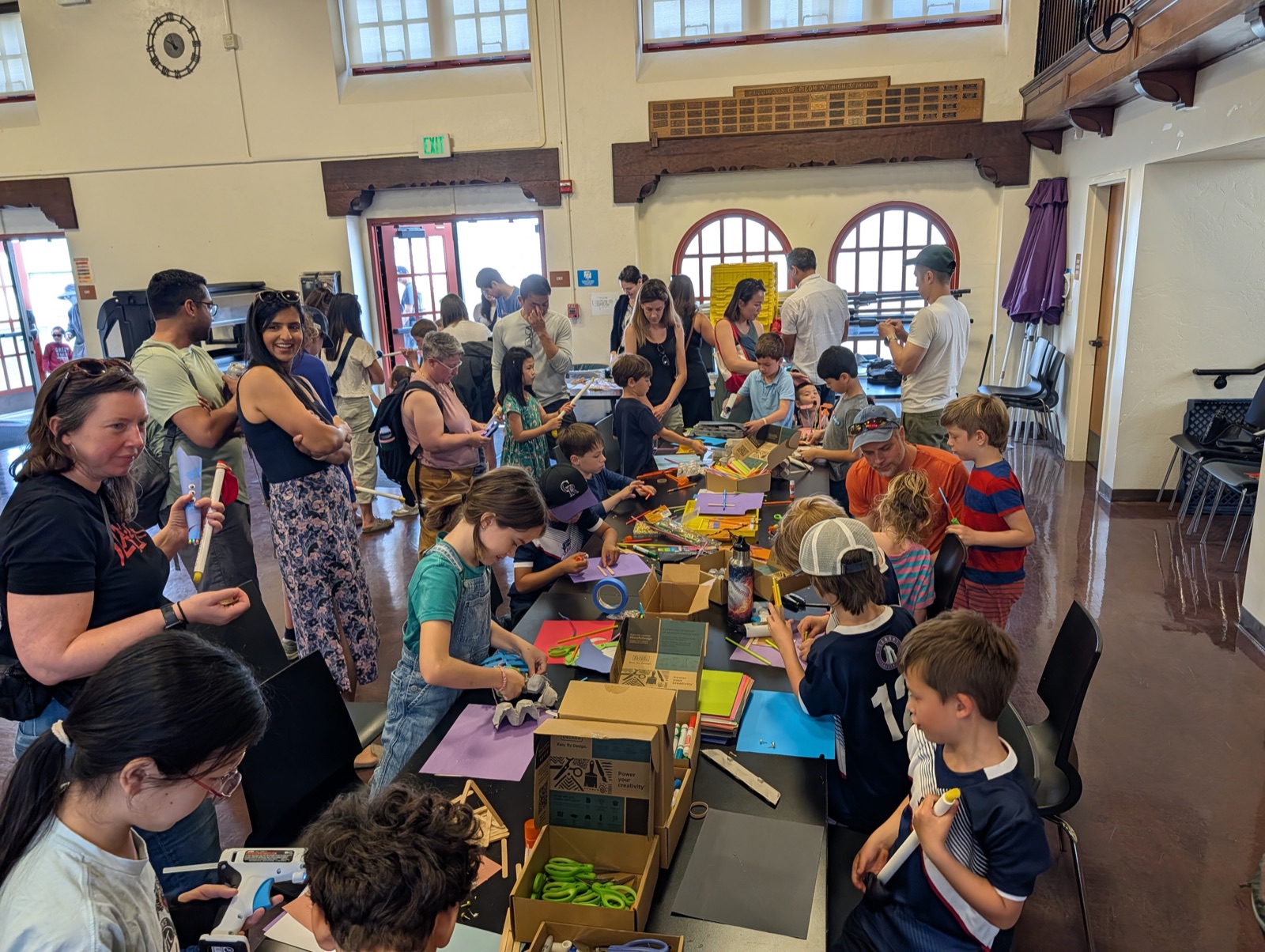 Children and parents at long tables making craft projects with paper, markers, and cardboard inside a community hall.