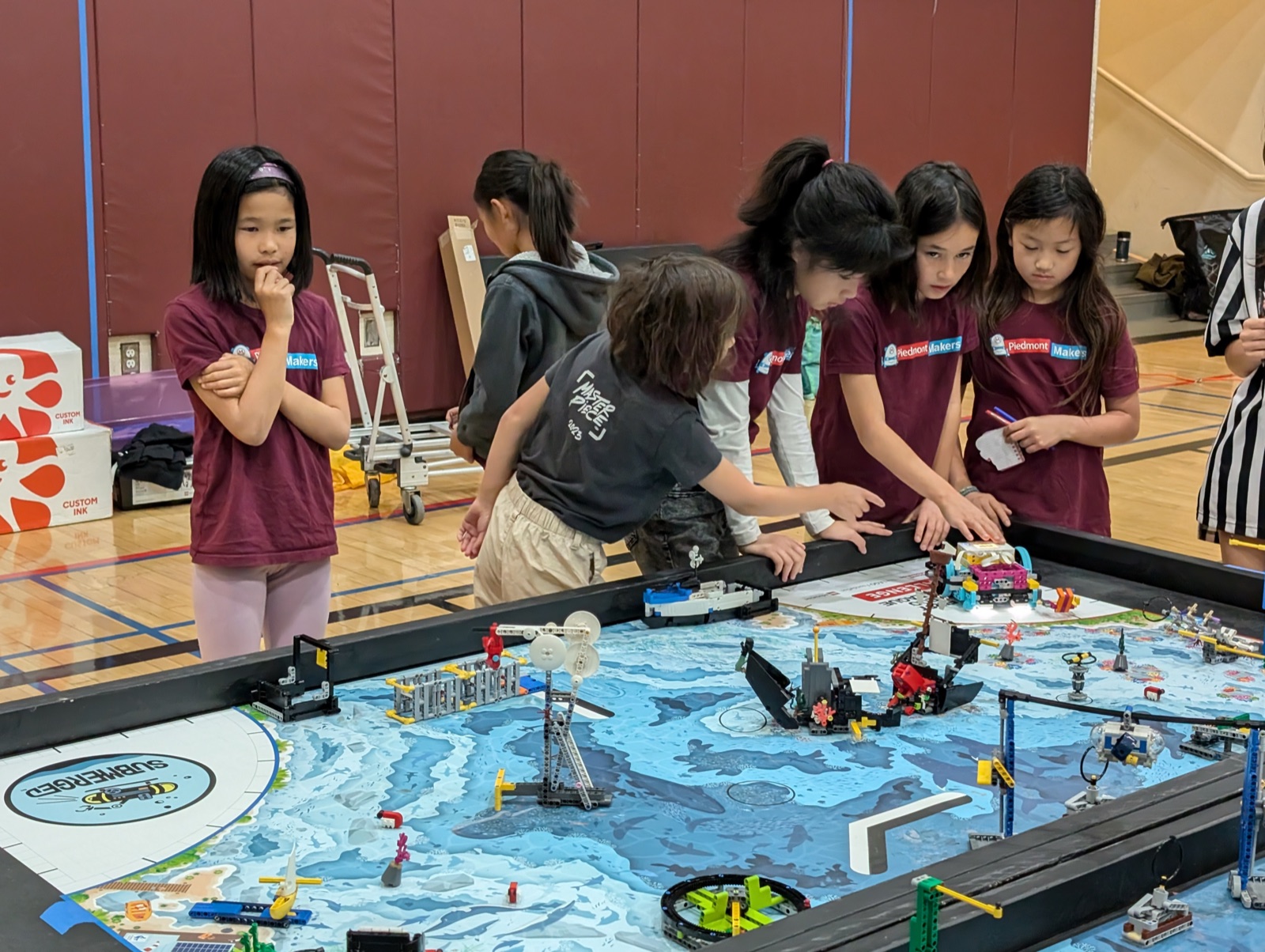 Five girls in maroon Piedmont Makers shirts working at an FLL Challenge competition table with a LEGO ocean-themed game board.