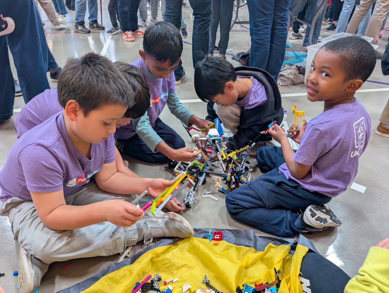 Four young boys in purple Piedmont Makers shirts sitting on the floor of a gym building LEGO Technic structures.