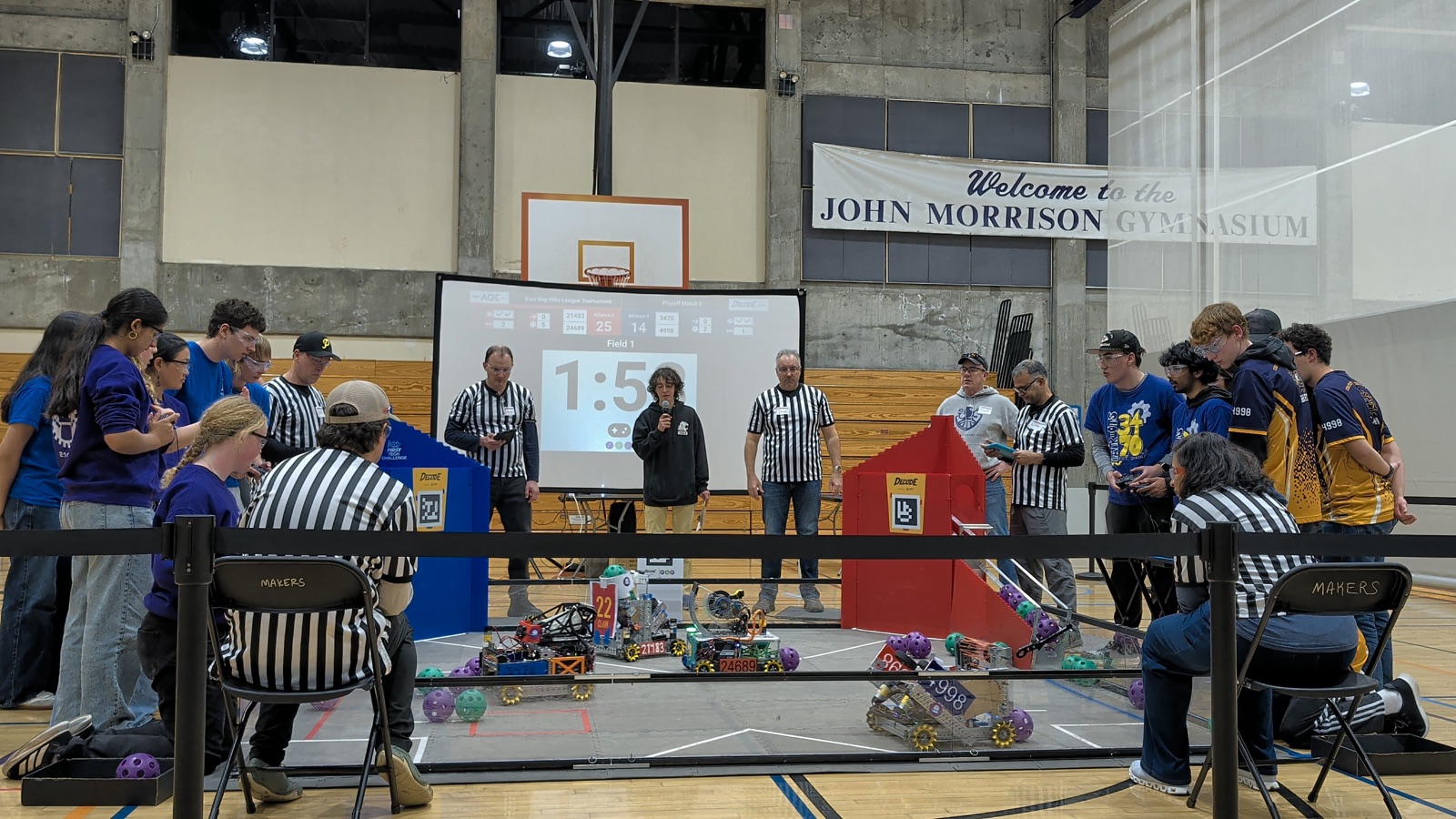FTC playoff match in progress at John Morrison Gymnasium — referees, team members at the alliance stations, and robots on the competition field.