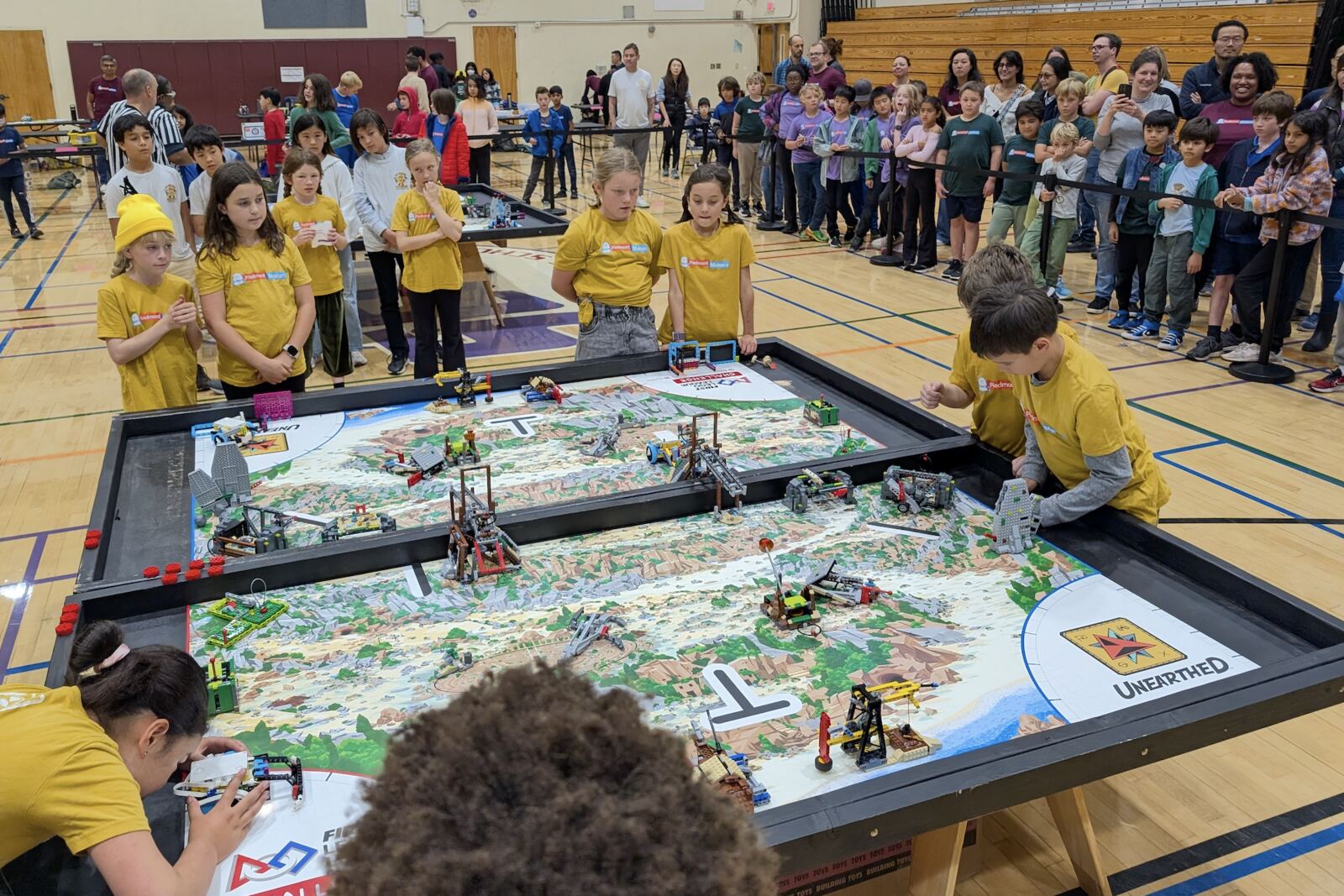 An FIRST LEGO League Challenge team in yellow Piedmont Makers shirts at the UNEARTHED-season tournament — kids around a large LEGO competition table with the game map laid out, big crowd of spectators behind.